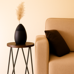 Black vase with dried pampas grass on a small table next to a beige couch with a brown pillow.