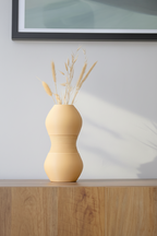 Beige vase with dried plants on a wooden surface against a light gray wall.